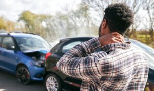 A young man rubbing his neck in pain from a whiplash injury, standing beside a damaged car after a traffic accident.