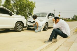 Asian man and African American woman sitting in a car, looking stressed after being involved in a serious accident. They are waiting for the insurance company to assess the damage to the vehicle's front bumper